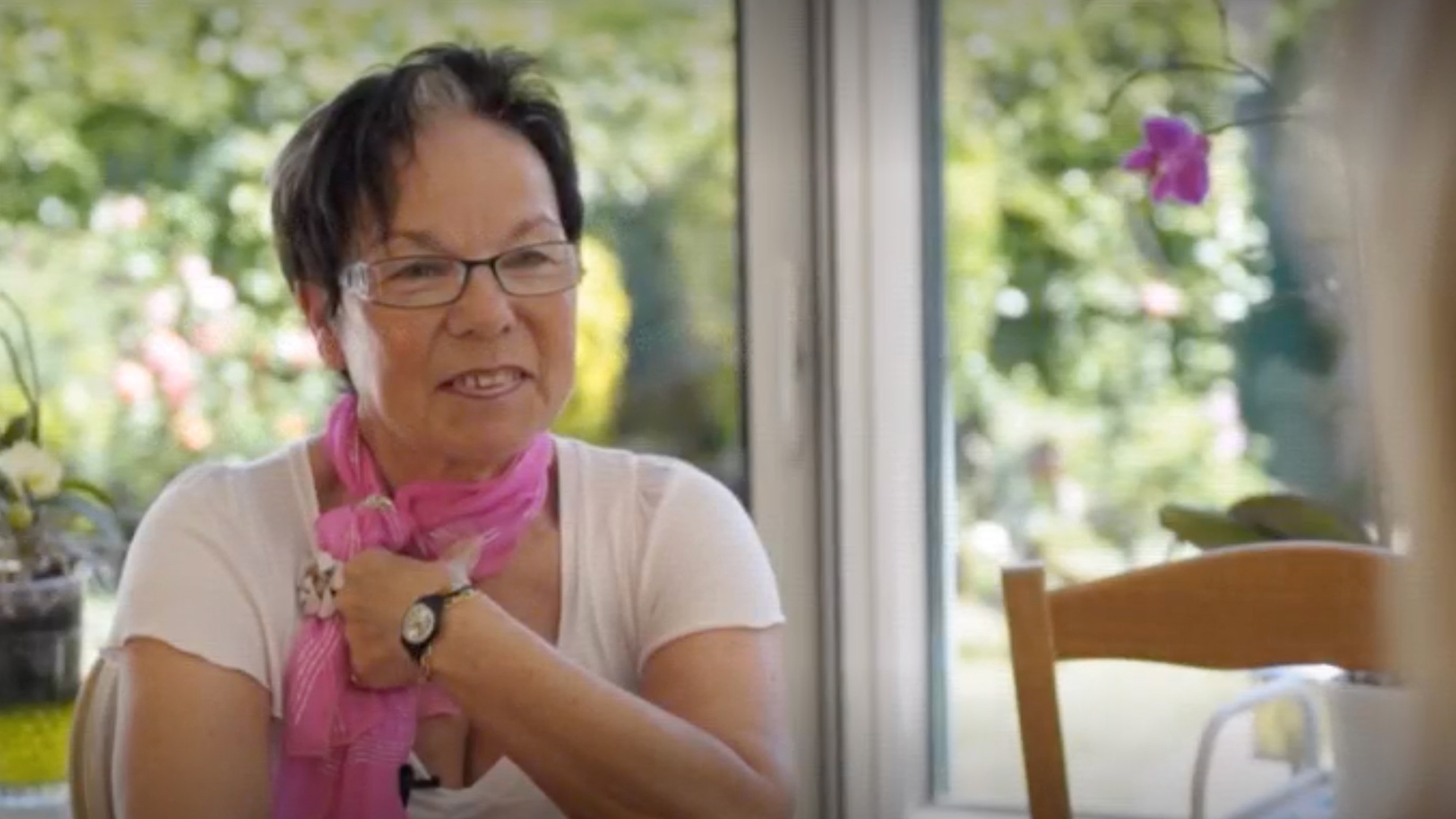 Image of Noelle Klein, a woman with a laryngectomy. She is smiling, wearing a pink scarf and pressing the device in her neckstoma.