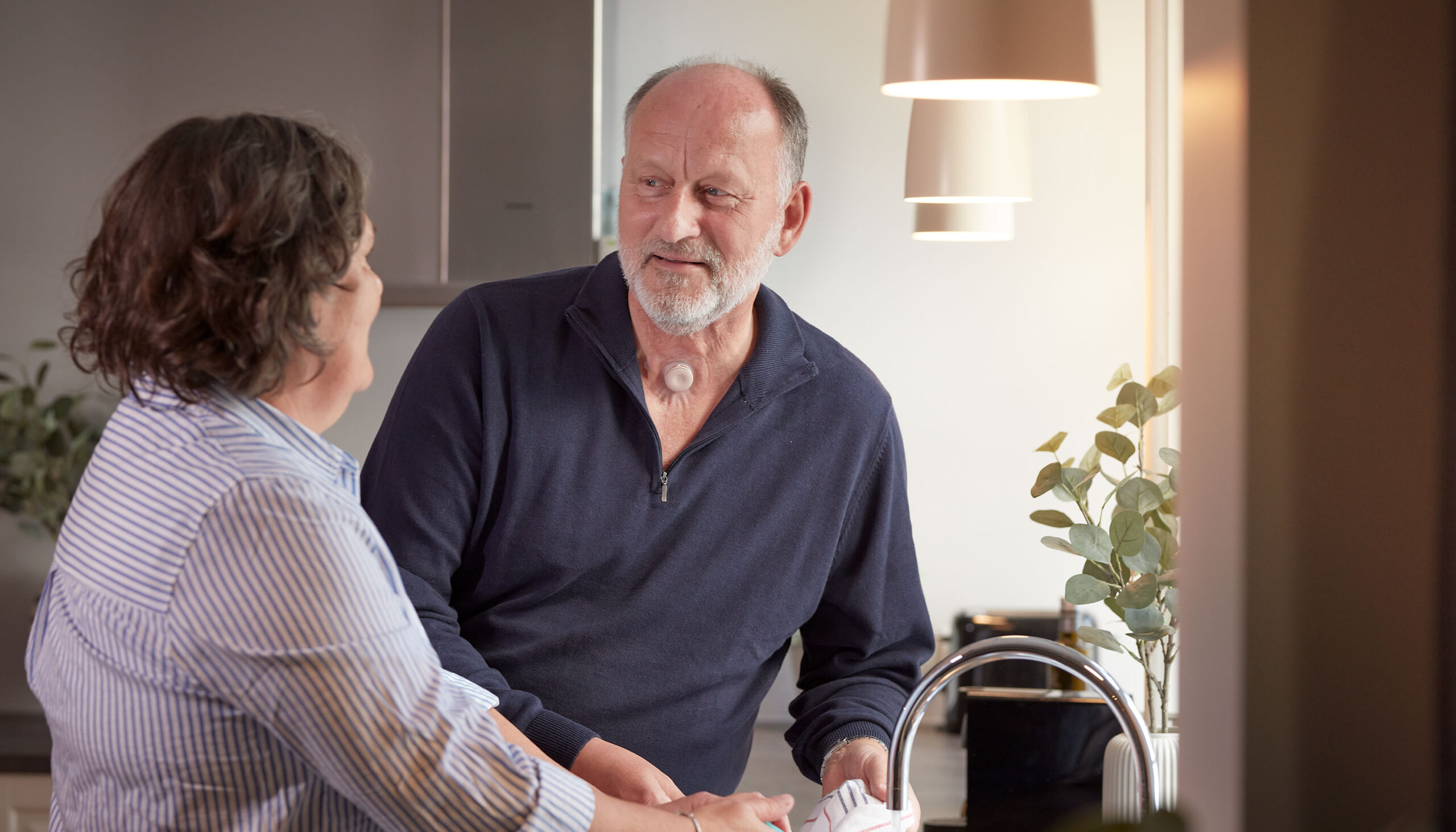 Man with a neck stoma helping woman to wash dishes while using a Provox life home HME